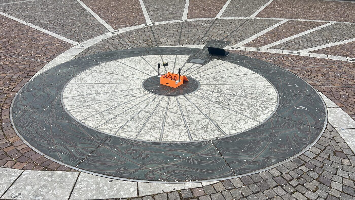 An orange 3D-printed box with two antennas sitting on the dark centre dot of a large painted disc in a paved square.