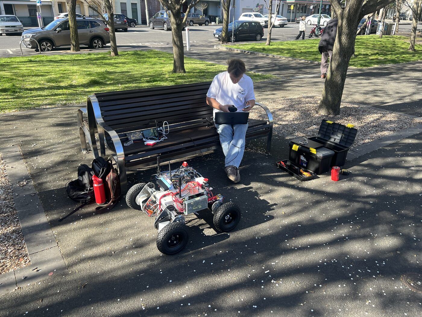 A team member on a park bench with a laptop and the base station, Prototype 2 parked on the path in front, with toolboxes and a fire extinguisher beside the bench.