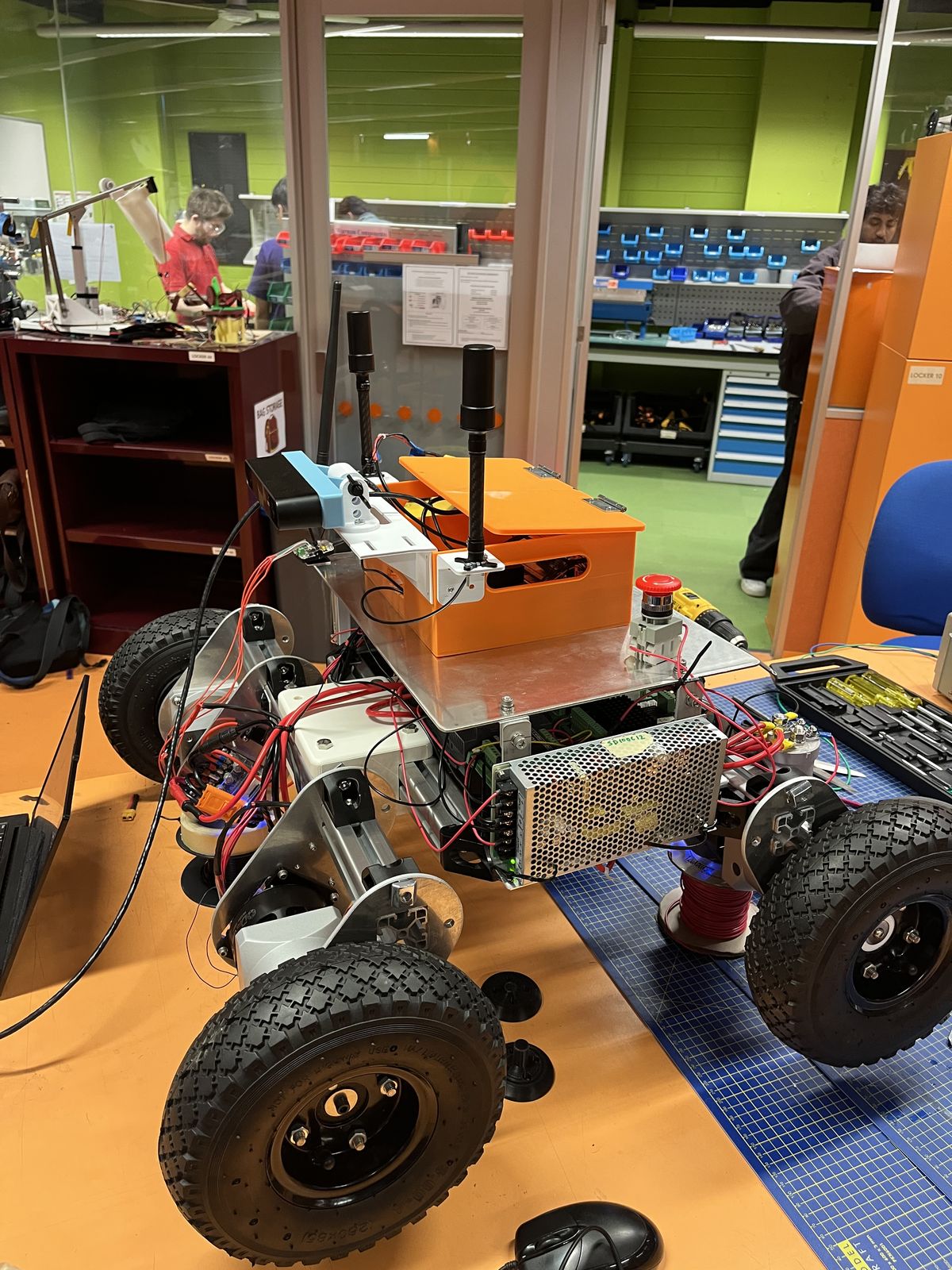 Prototype 2 on a workbench with an orange enclosure on top, two antenna masts, a stereo camera, a red E-stop button and a power supply on the side.