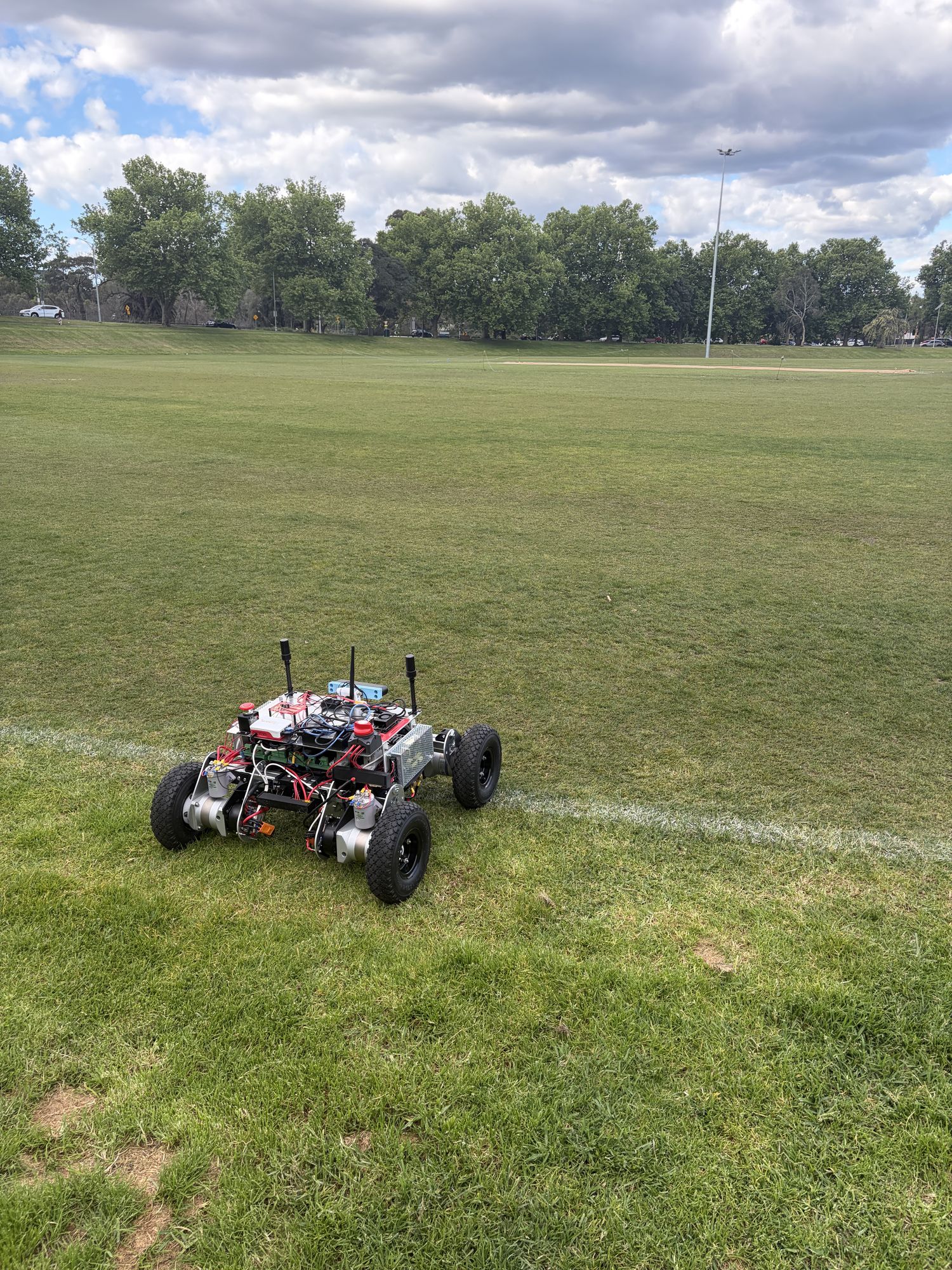 Prototype 2, a four-wheeled aluminium rover with three antenna masts, sitting on a mown grass oval beside a painted line, with trees and cloudy sky behind.