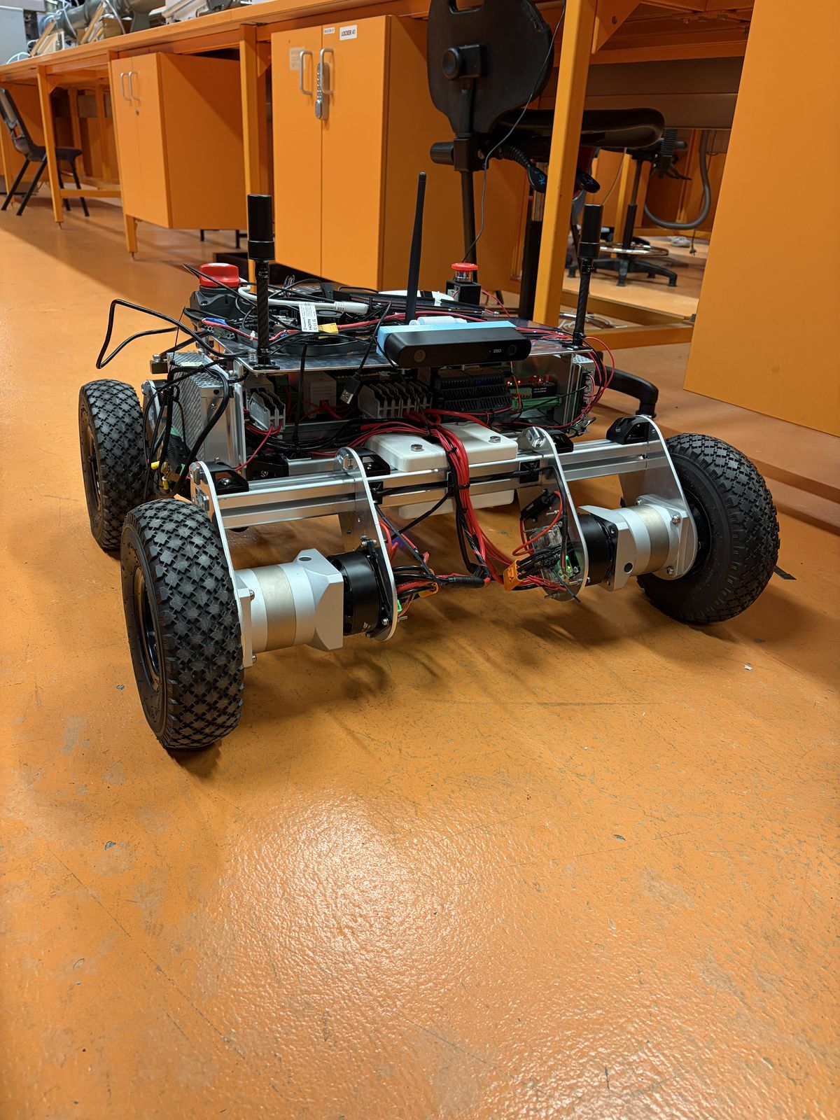 Prototype 2 on the lab floor with its electronics fitted: two GNSS antenna masts, a stereo camera on top, red emergency-stop buttons and bundles of red and black wiring.